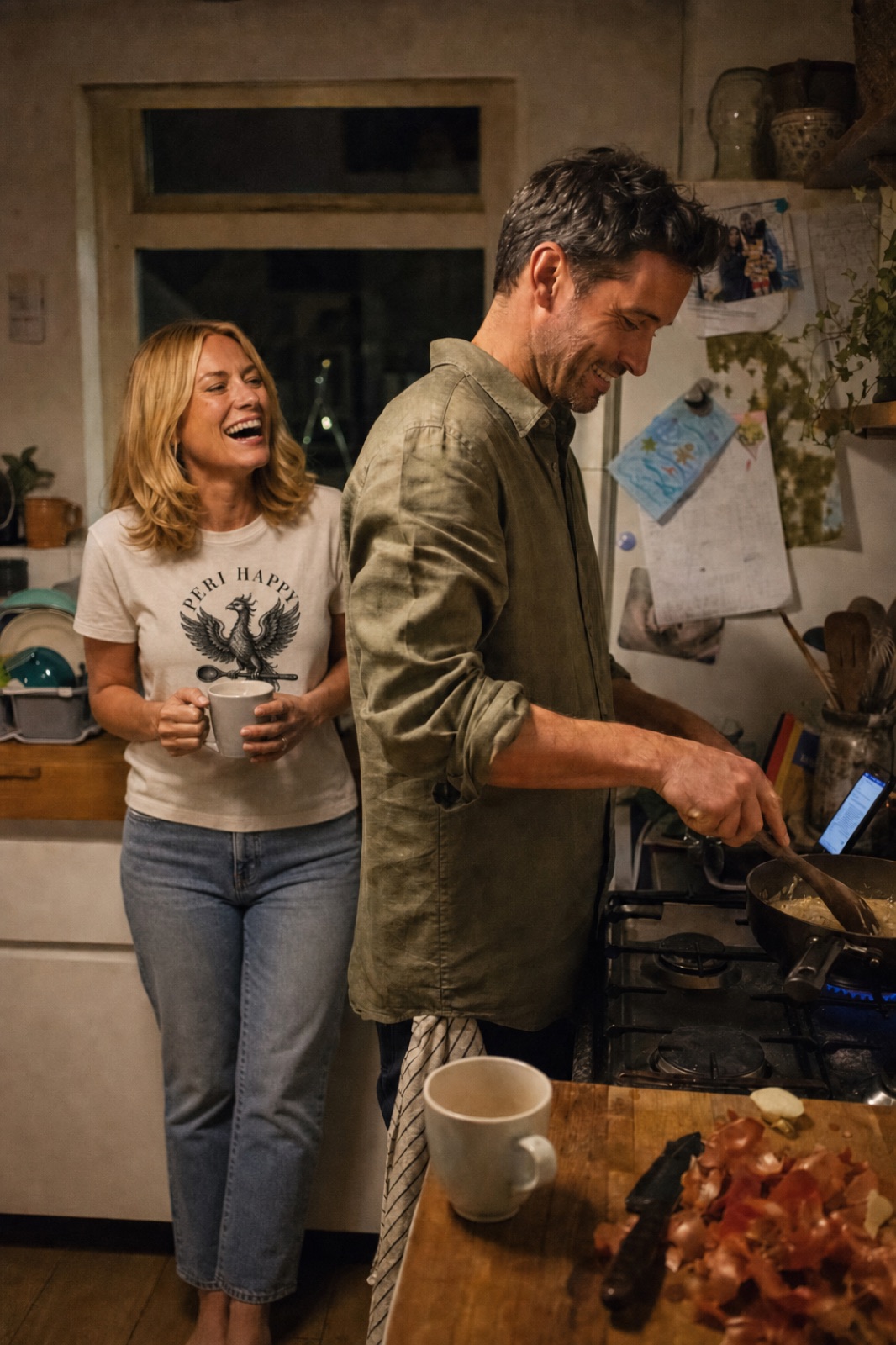 A candid weeknight moment: a man cooks at the hob while his partner, wearing the ecru Phoenix Tee, leans on the counter with a mug of tea, laughing at something he said